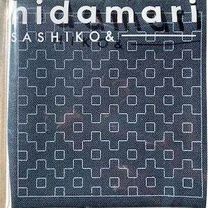 May include: A dark blue fabric square featuring a repeating white geometric pattern of interlocking crosses and squares. The text "hidamari" and "SASHIKO &" is printed in white across the top portion of the material, which is encased in clear plastic.