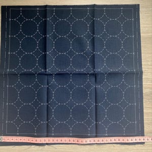 Könnte beinhalten: Ein marineblaues quadratisches Gewebe mit einem weiß genähten geometrischen Muster aus Kreisen und Linien. Der Stoff hat einen weißen Rand und wird mit einem rosa Maßband gemessen. Der Stoff ist etwa 91 cm lang.