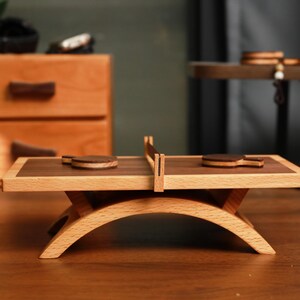 May include: A miniature wooden ping pong table with two paddles and a net. The table has a light wood frame with a dark wood playing surface. The table is supported by a curved, light wood base. Other wooden objects are in the background.