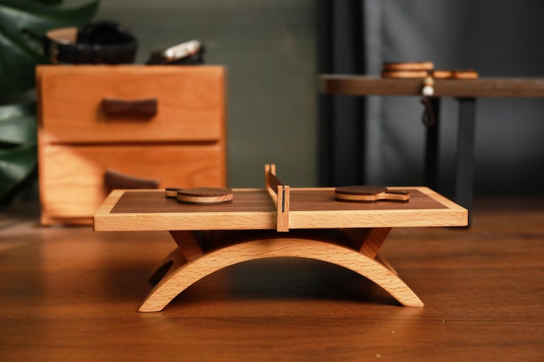 May include: A miniature wooden ping pong table with two paddles and a net. The table has a light wood frame with a dark wood playing surface. The table is supported by a curved, light wood base. Other wooden objects are in the background.