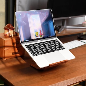 以下が含まれることがあります： 黒いキーボードが付いたシルバーのラップトップが木製のスタンドに置かれています。画面には漫画のシーンが表示されています。左側には、2つの引き出しとキノコの置物が入った小さな木箱があります。机は濃い色の木でできています。