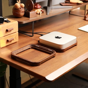 May include: A wooden desk with a light-coloured computer and a matching wooden frame. A small wooden drawer unit with two drawers and a watch are on the left. Decorative items are on a shelf above.