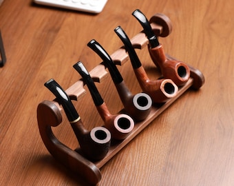 May include: A wooden pipe rack holding five tobacco pipes. The pipes have dark stems and light-colored bowls. The rack is made of wood and has a curved design on the sides. The pipes are arranged in a row, with the bowls facing forward.