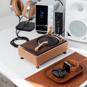 May include: A wooden turntable with a black record player on top. The turntable is sitting on a white desk with a brown leather mat. There is a small wooden figurine of a cat standing on the turntable.