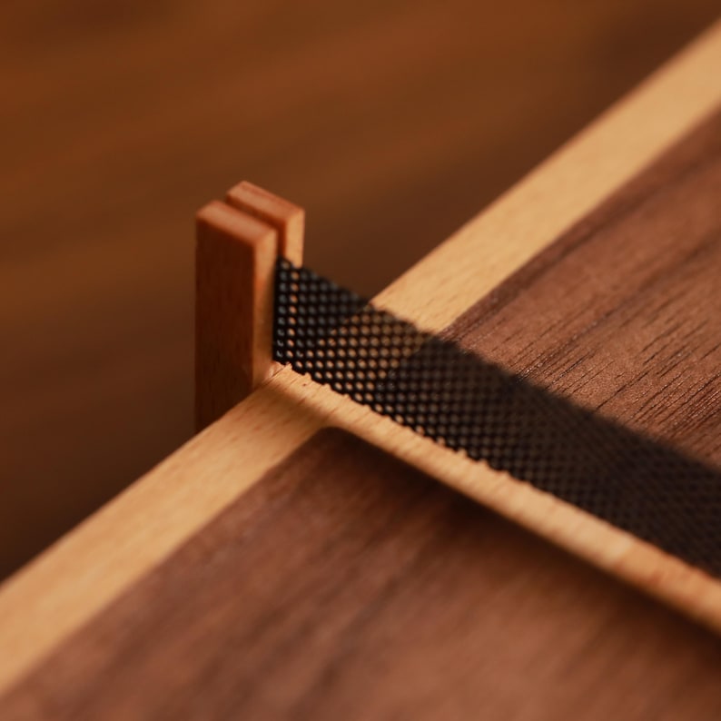 May include: Close-up of a wooden box with a dark brown finish and light brown trim. A black mesh band is attached to the side of the box. The box is made of wood and has a minimalist design.