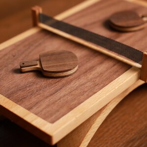 May include: A wooden ping pong set with a dark brown playing surface and light brown frame. The set includes two small paddles and a net. The paddles are made of layered wood. The net is black and mesh.