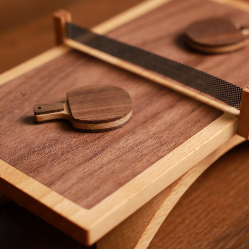 May include: A wooden ping pong set with a dark brown playing surface and light brown frame. The set includes two small paddles and a net. The paddles are made of layered wood. The net is black and mesh.