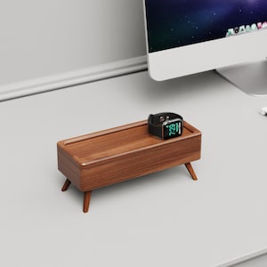 Puede incluir: Un soporte de reloj de madera con un reloj en la parte superior. El soporte tiene una base rectangular con bordes redondeados y cuatro patas. El reloj es negro con una pantalla digital que muestra la hora 7:29.