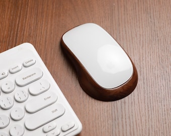 May include: A white wireless computer mouse with a wooden base sits on a wooden surface next to a white keyboard. The mouse has a smooth, glossy white top and a dark brown wooden base. The keyboard has white keys.
