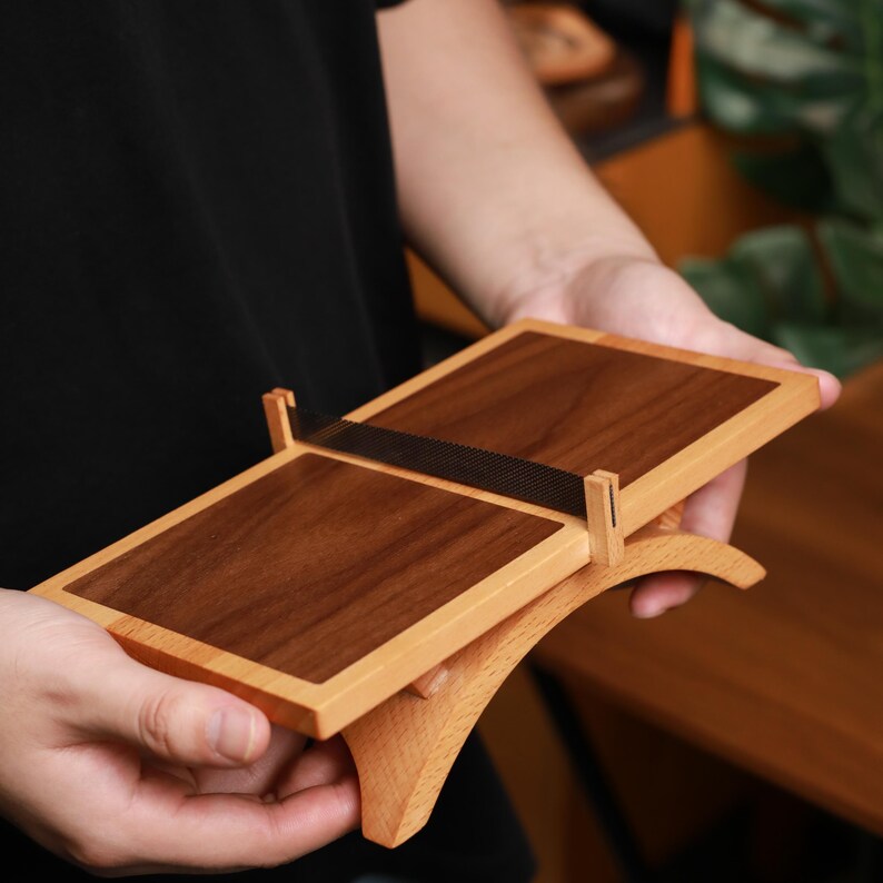 May include: A wooden table tennis net set, featuring a rectangular design with a dark brown playing surface and a lighter wood frame. A black mesh net is stretched across the top. The set is held in someone's hands.