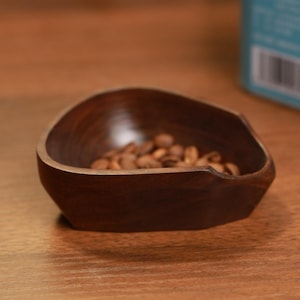 Könnte beinhalten: Eine kleine, dunkelbraune Holzschale gefüllt mit gerösteten Kaffeebohnen. Die Schale hat eine einzigartige, asymmetrische Form und steht auf einer Holzoberfläche. Die Kaffeebohnen sind hellbraun, und die Holzmaserung der Schale ist sichtbar.