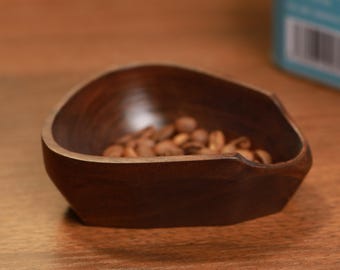 May include: A small, dark brown wooden bowl containing roasted coffee beans. The bowl has an unusual, asymmetrical shape and rests on a wooden surface. The coffee beans are light brown, and the wood grain of the bowl is visible.