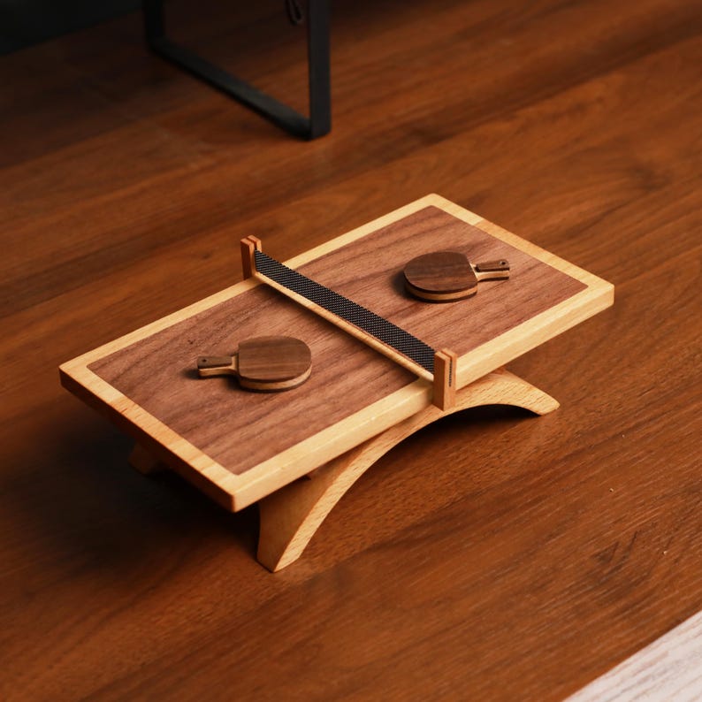 May include: A wooden ping pong game set. The set includes a rectangular playing surface with a dark wood center and light wood border, two small paddles, and a net. The set is elevated on a curved wooden base.