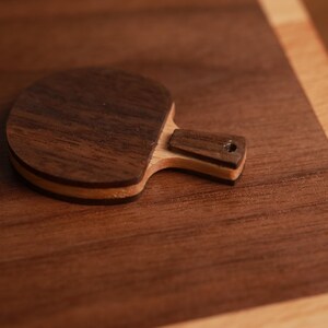 May include: A small, wooden ping pong paddle with a dark brown face and a lighter brown handle. The paddle is made of layered wood, with a light-colored layer visible around the edge. The handle has a small hole.