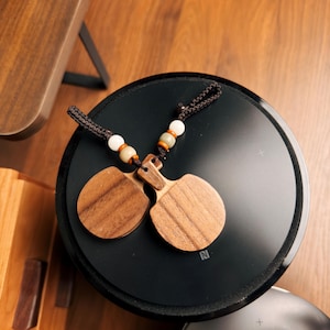 Könnte beinhalten: Zwei Holz-Kastagnetten mit einer dunkelbraunen Kordel und dekorativen Perlen liegen auf einer schwarzen Oberfläche. Die Kastagnetten sind aus hellbraunem Holz mit sichtbarer Maserung gefertigt. Die Perlen sind weiß, beige und orange. Die schwarze Oberfläche hat ein silbernes "N"-Logo.