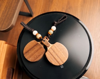 Op de afbeelding: Twee houten castagnetten met een donkerbruin koord en decoratieve kralen liggen op een zwart oppervlak. De castagnetten zijn gemaakt van lichtbruin hout met zichtbare nerf. De kralen zijn wit, beige en oranje. Het zwarte oppervlak heeft een zilveren "N"-logo.