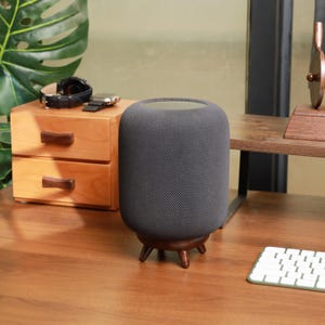 Puede incluir: Un altavoz inteligente cilíndrico gris oscuro con una cubierta de tela, que descansa sobre una base de madera con tres patas. El altavoz está sobre un escritorio de madera, con una pequeña unidad de cajones de madera y un teclado en el fondo.