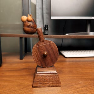 May include: A wooden ping pong paddle with a dark brown finish, resting on a matching wooden stand. The paddle features a small, round ball in the center. The design is minimalist and elegant, suitable for home decor or a desk accessory.