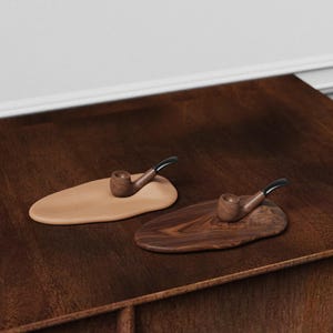 May include: Two wooden tobacco pipes rest on individual, organic-shaped stands. One stand is a light tan color, while the other is a dark brown with a wood grain pattern. The pipes have a dark finish and black stems.
