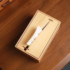 May include: A light-colored wooden tissue box with a rectangular shape. The box has a slot on the top for tissue dispensing and a cross-shaped design on the front. White tissues are visible, partially pulled from the opening.