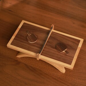 May include: A wooden ping pong-themed display. The display features two rectangular wooden surfaces with a dark brown finish, each with a small ping pong paddle. A net divides the two surfaces. The display is supported by a light-colored wooden base.