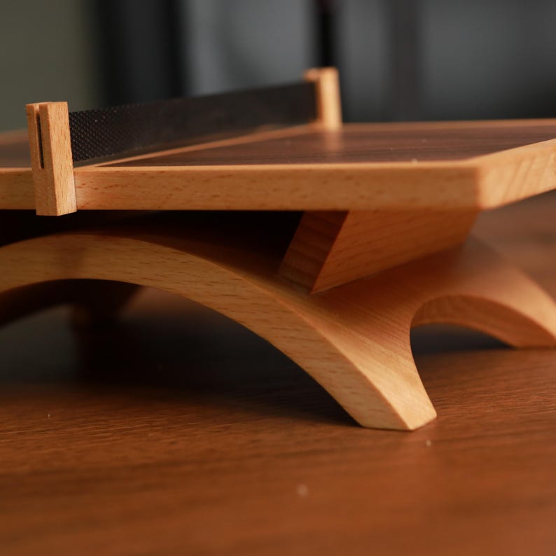 May include: A small, wooden table with a dark brown tabletop and light brown legs. The legs have a curved design. A black mesh net is stretched across the top. The table is made of light-colored wood.
