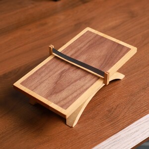 May include: A wooden book stand with a rectangular design. The stand features a dark brown, rectangular surface framed by a lighter wood border. A black strap secures the book. The stand is supported by a curved base.