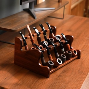 May include: A wooden pipe rack displaying several tobacco pipes. The pipes have dark stems and various bowl shapes, with a mix of light and dark wood tones. The rack is made of dark wood and holds the pipes upright.