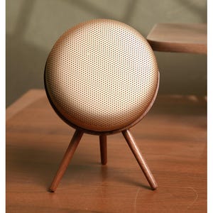 May include: A gold-colored, round speaker with a perforated surface sits on a wooden tripod stand. The speaker has a dark wooden frame and the tripod legs are also made of wood. The speaker is on a wooden surface.