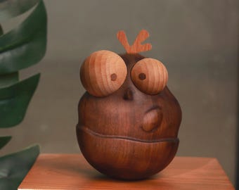 May include: A whimsical wooden sculpture of a creature with large, round eyes and a small, curved mouth. The sculpture is made of dark brown wood with lighter wood accents for the eyes and a small detail on top. It sits on a wooden surface.
