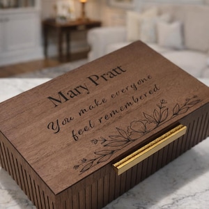 May include: A rectangular dark brown wooden box with a hinged lid featuring a gold-toned metal handle. The lid is engraved with the text Mary Pratt, You make everyone feel remembered, and a floral line art design on the right side.