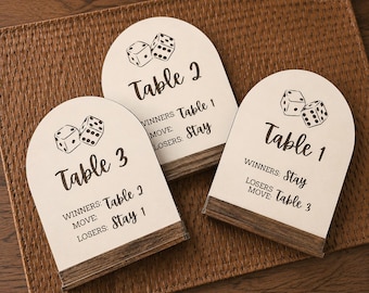 May include: Three arched off-white table signs with dark wood bases. Each sign features a black graphic of two dice and text indicating table numbers, winners, and loser moves. The text reads Table 1, Table 2, and Table 3 with specific game instructions.