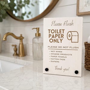 Puede incluir: Un letrero rectangular de madera clara con texto en marrón oscuro que dice: Please Flush TOILET PAPER ONLY. PLEASE DO NOT FLUSH: WET WIPES, HYGIENE PRODUCTS, PAPER TOWELS, COTTON PADS, DIAPERS. Thank you! El letrero presenta dos pequeños detalles circulares negros.