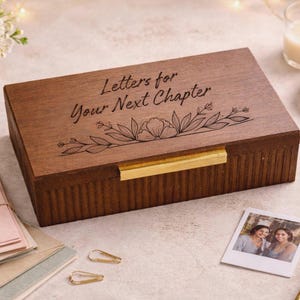 May include: A rectangular dark brown wooden box with a ribbed side texture and a gold metal latch. The lid is engraved with the text "Letters for Your Next Chapter" above a line drawing of a floral vine. The box is a storage container for items.