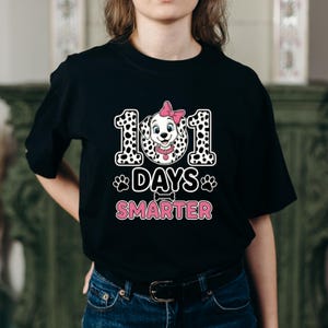 K&ouml;nnte beinhalten: Schwarzes T-Shirt mit einem Dalmatiner-Hund-Motiv mit rosa Schleife, umgeben von den Zahlen "101" im Dalmatiner-Print. Der Text "101 Days Smarter" wird darunter in Rosa angezeigt.