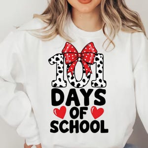 K&ouml;nnte beinhalten: Wei&szlig;es Sweatshirt mit einem Grafikdesign, das 101 Schultage feiert. Das Design zeigt die Zahl 101 in einem schwarz-wei&szlig;en Dalmatiner-Print, eine rote Schleife mit wei&szlig;en Punkten und die Worte "DAYS OF SCHOOL" mit roten Herzen.