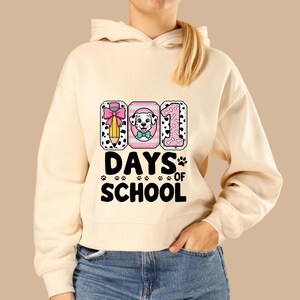 K&ouml;nnte beinhalten: Cremefarbener Kapuzenpullover mit Grafikdesign. Das Design zeigt den Text "101 DAYS OF SCHOOL" mit einem Bleistift, einem Dalmatiner und der Zahl 1. Das Design enth&auml;lt Pfotenabdr&uuml;cke und eine rosa Schleife.