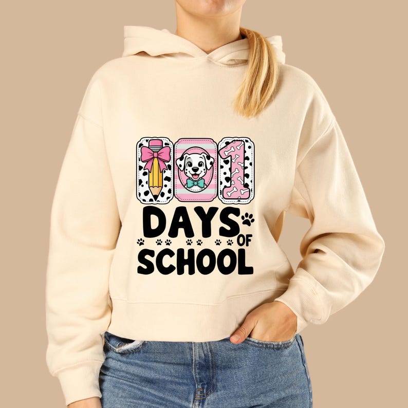 K&ouml;nnte beinhalten: Cremefarbener Kapuzenpullover mit Grafikdesign. Das Design zeigt den Text "101 DAYS OF SCHOOL" mit einem Bleistift, einem Dalmatiner und der Zahl 1. Das Design enth&auml;lt Pfotenabdr&uuml;cke und eine rosa Schleife.
