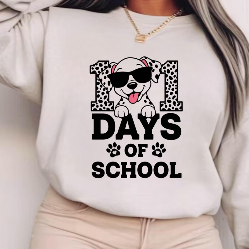K&ouml;nnte beinhalten: Ein cremefarbenes Sweatshirt mit einer Grafik, die einen Dalmatinerhund mit Sonnenbrille, die Zahl 101 und den Text "DAYS OF SCHOOL" in Schwarz zeigt. Der Hund hat eine rosa Zunge herausgestreckt.