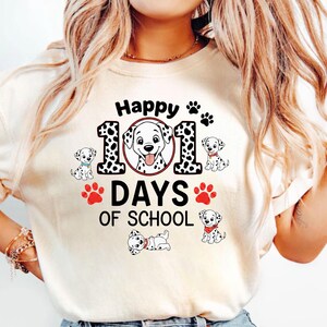 K&ouml;nnte beinhalten: Cremefarbenes T-Shirt mit einem Grafikdesign, das einen Dalmatinerhund und den Text "Happy 101 Days of School" zeigt. Das Design beinhaltet Pfotenabdr&uuml;cke und Dalmatiner-Welpen. Das Shirt ist ein Freizeitartikel.