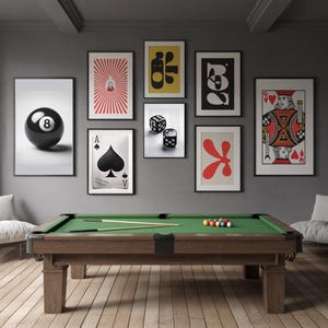 May include: A green felt pool table with a brown wooden frame and black legs. The table is set up for a game of pool with balls and cues. The table is in a room with a gray wall and several framed prints on the wall. The prints feature a black and white playing card, a red and white playing card, a black and white 8-ball, a red and white abstract design, a yellow and black abstract design, a black and white abstract design, and a red and white abstract design.