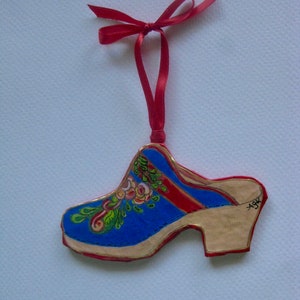 May include: A blue and gold painted wooden clog ornament with red and white floral details. The ornament is attached to a red ribbon.
