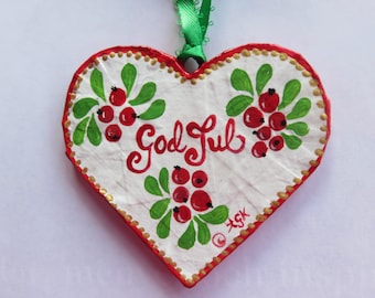 May include: Heart-shaped Christmas ornament with a white background, red trim, and gold accents. The ornament features green leaves, red berries, and the words "God Jul" in red script. A green ribbon is attached for hanging.