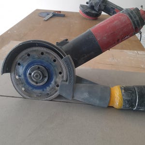 May include: A close-up of a black and red angle grinder with a diamond blade attached. The grinder is resting on a piece of wood and has a yellow and black dust collection hose attached.