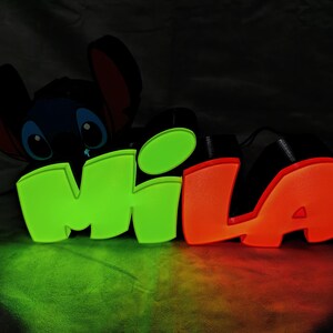 Puede incluir: Letrero de neón brillante con el nombre "MILA" en verde y naranja, con un personaje de dibujos animados de Stitch. El letrero es negro con un brillo de neón, ideal para la habitación de un niño. La iluminación es verde y naranja.
