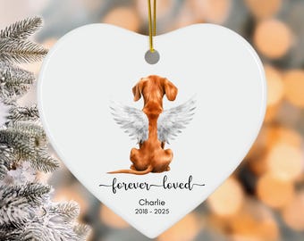 May include: A white heart-shaped ornament with a golden string for hanging. It features a brown dog with white angel wings, the words "forever loved", and the name "Charlie" with the years 2018-2025.