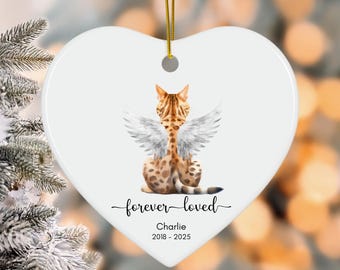 May include: White heart-shaped ornament with a Bengal cat illustration wearing angel wings. The ornament has the text "forever loved" and the name "Charlie" with dates 2018-2025. A gold string is attached for hanging.