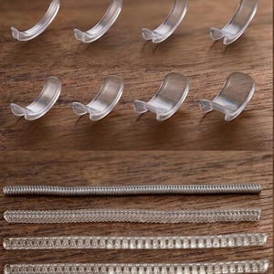 Op de afbeelding: Een verzameling doorzichtige plastic clips en bindruggen. De clips zijn gebogen met een kleine lip, en de ruggen zijn er in verschillende stijlen, waaronder een metalen spiraal en verschillende doorzichtige plastic strips met in elkaar grijpende tanden. De items zijn gerangschikt op een houten oppervlak.