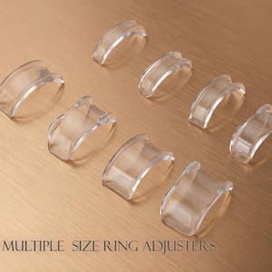 Puede incluir: Ocho ajustadores de anillos de plástico transparente están dispuestos sobre una superficie marrón claro. Los ajustadores son curvos y tienen una ranura para adaptarse a una banda de anillo. El texto en la parte inferior dice "8 MULTIPLE SIZE RING ADJUSTERS."