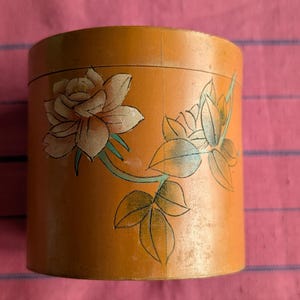 Puede incluir: Un recipiente redondo de madera con un diseño floral. El recipiente está pintado en tonos de naranja, marrón, verde y blanco. El diseño presenta una rosa grande y hojas.